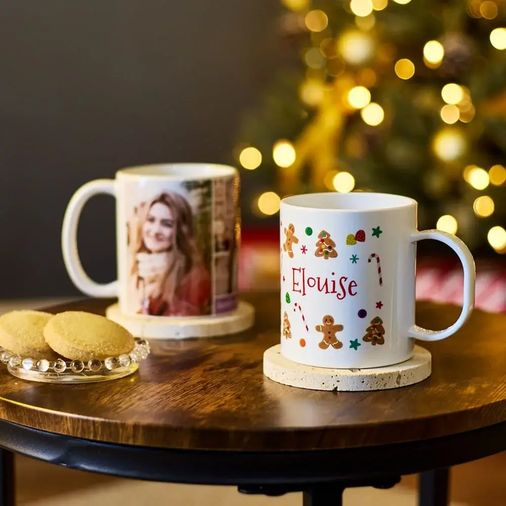 Two personalised mugs on a wooden table, one decorated with festive gingerbread and candy cane designs featuring the name Eloise, accompanied by a small plate of cookies, with a blurred Christmas tree in the background.